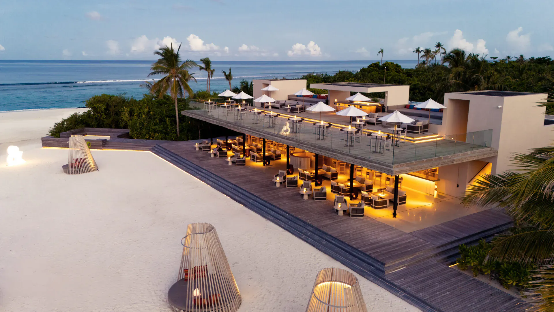 Aerial view of RAAYA by Atmosphere beachfront bar at dusk in Maldives, with palm trees, ocean, and lit seating.