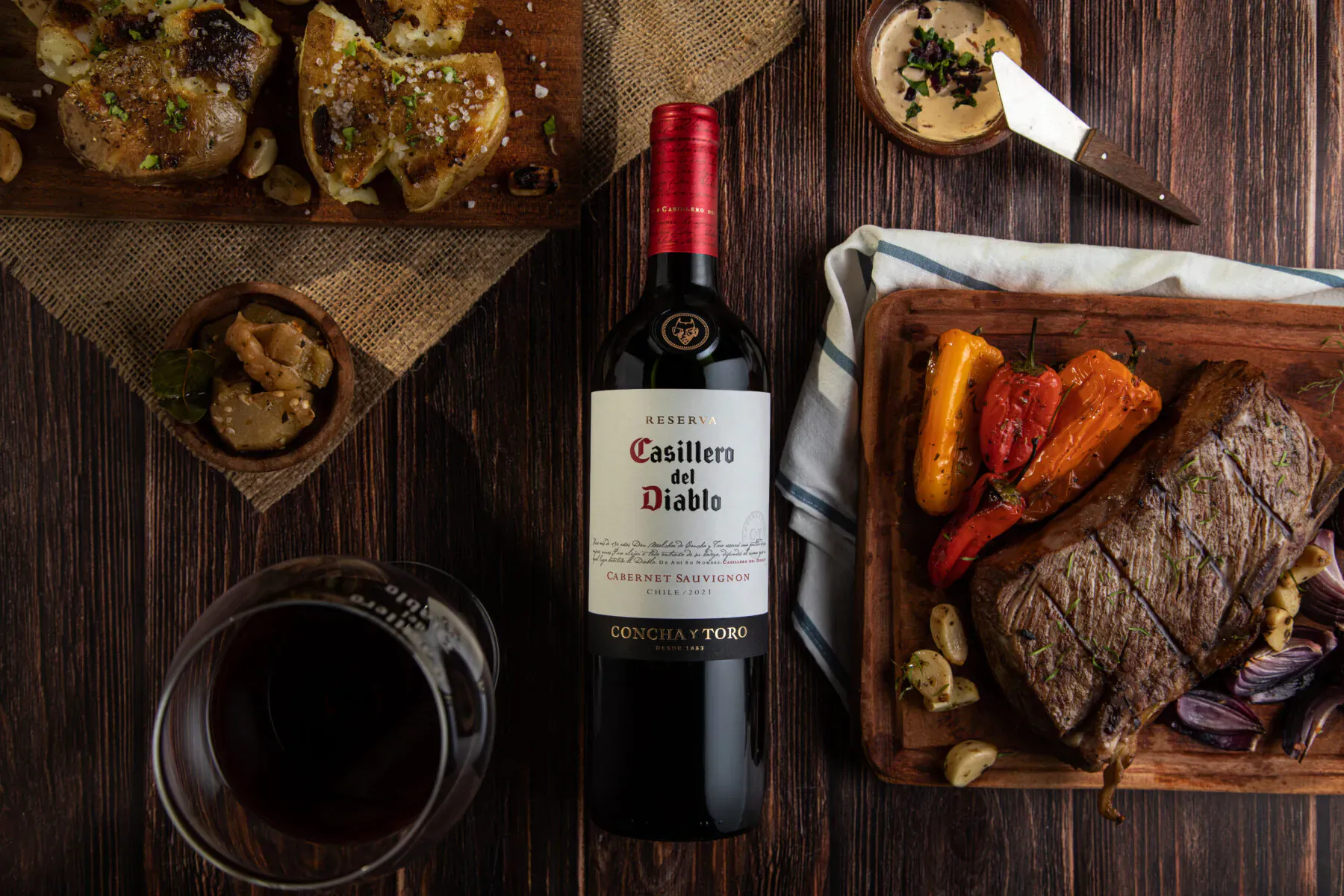 Casillero del Diablo red wine bottle with grilled steak, roasted peppers, mushrooms on wooden board, wine glass on table.