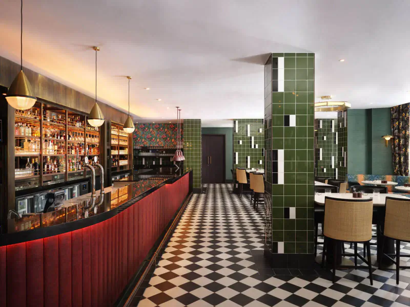Art deco-style bar at The Other House with green tiled pillars, brass shelving stocked with liquor, red curved counter, black-and-white checkered floor, and dining tables.