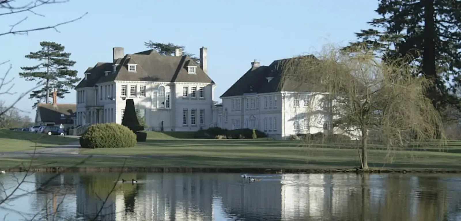 Brockencote Hall Hotel: luxury white manor house with brown roofs by lake, reflected in water amid trees and lawns.