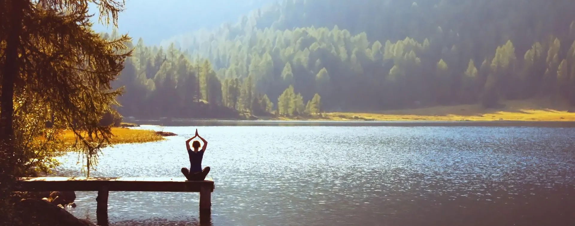 Person in lotus yoga pose with hands raised on wooden pier over serene mountain lake at sunset