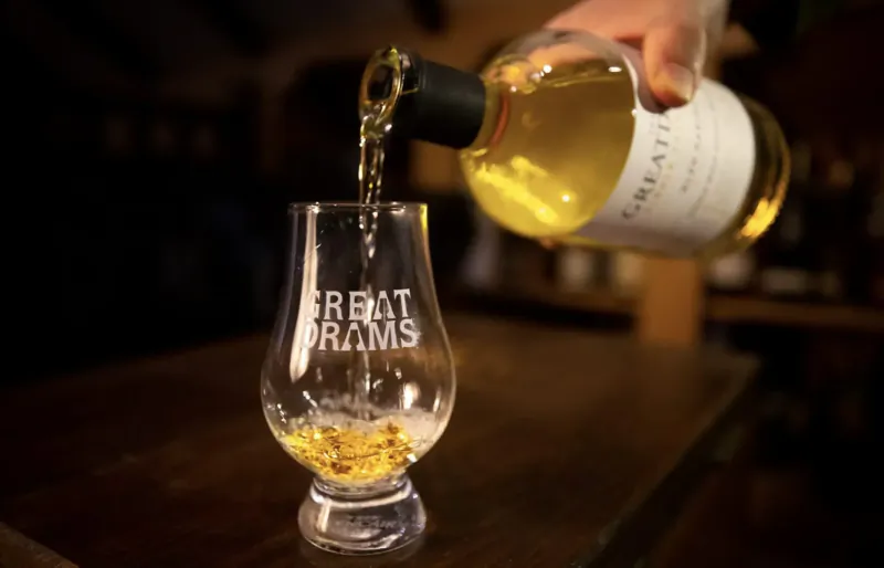 Hand pouring GreatDrams whisky from bottle into etched 'Great Drams' Glencairn glass on wooden bar.