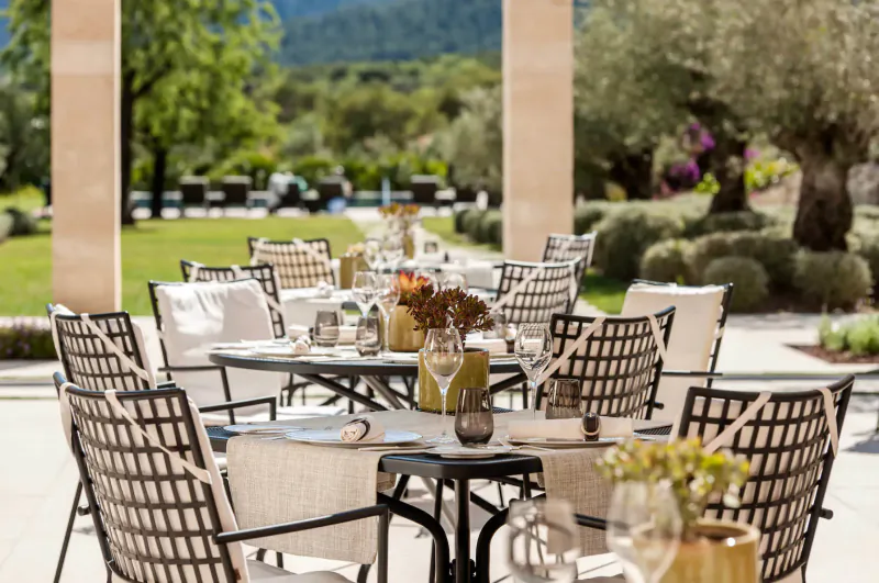 Elegant outdoor dining table set with glassware, flowers, and linens at Castell Son Claret, Mallorca, with olive trees and mountains.