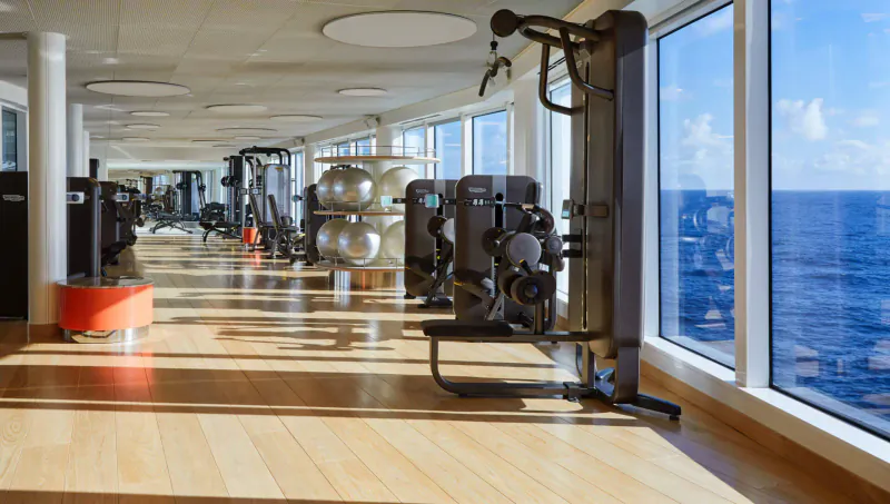 Spacious gym on Virgin Cruises ship with exercise machines, yoga balls, and ocean views through large windows.