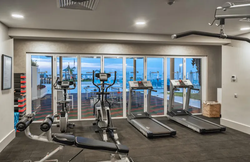 Modern hotel gym with treadmills, exercise bikes, weights, and pool view through large windows.