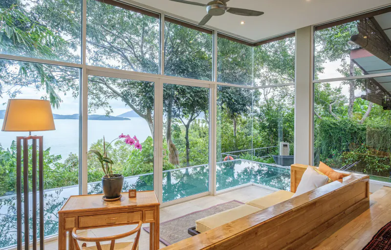 Spacious wellness retreat room with wooden desk, sofa, orchids, pool, and panoramic sea views through large windows.