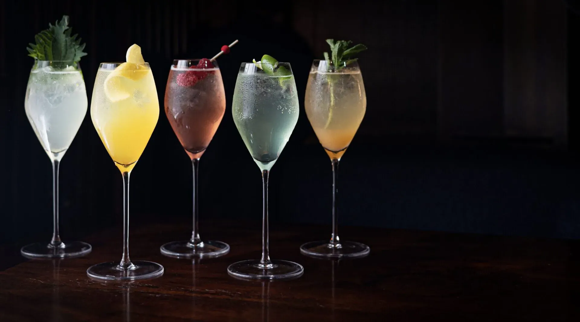 Four elegant cocktails garnished with lemon, cherry, lime, and mint on a dark table, Lucky Cat by Gordon Ramsay.