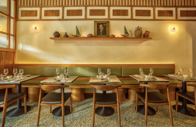 Elegant wooden restaurant booth with green cushions, set table for sharing, wall shelves with ceramics and portrait, warm lighting.