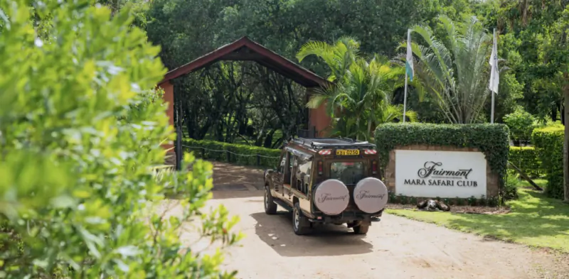 Fairmont Mara Safari Club entrance with wooden archway, lush greenery, flags, and Land Rover driving in.
