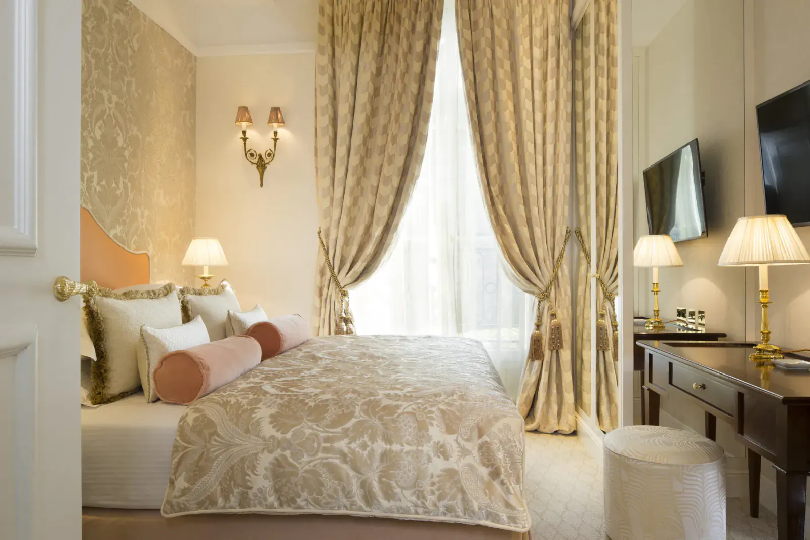Elegant luxury hotel bedroom with ornate beige bed, pillows, heavy curtains, lamps, TV, and desk at Relais Christine Paris.