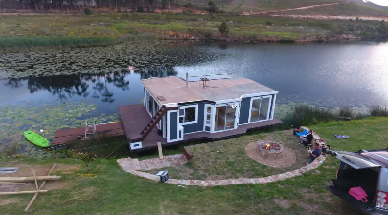 Aerial view of blue houseboat on lake with dock, kayak, campfire, people, and truck in grassy Elgin Valley setting.