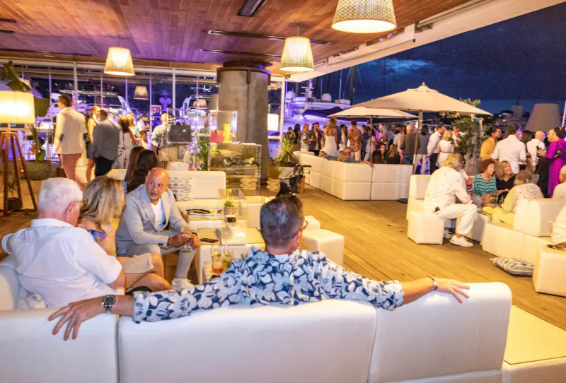 Lively outdoor evening event on wooden deck with white sofas, diverse guests chatting under pendant lamps, Balearic vibe.