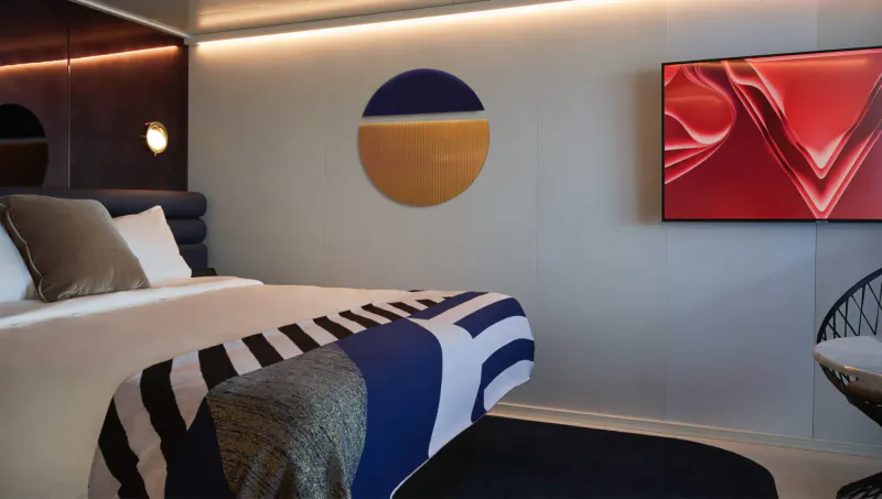 Luxurious adults-only cruise ship bedroom with modern bed, geometric wall art, red TV, and wicker chair.