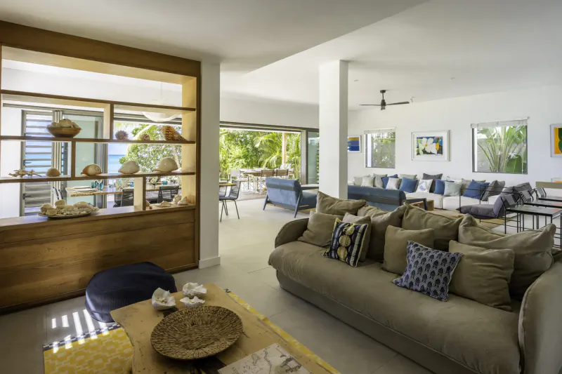 Spacious open-plan Villa Sapphire living room with wooden shelves of shells, beige sofas, tropical garden view through sliding doors.