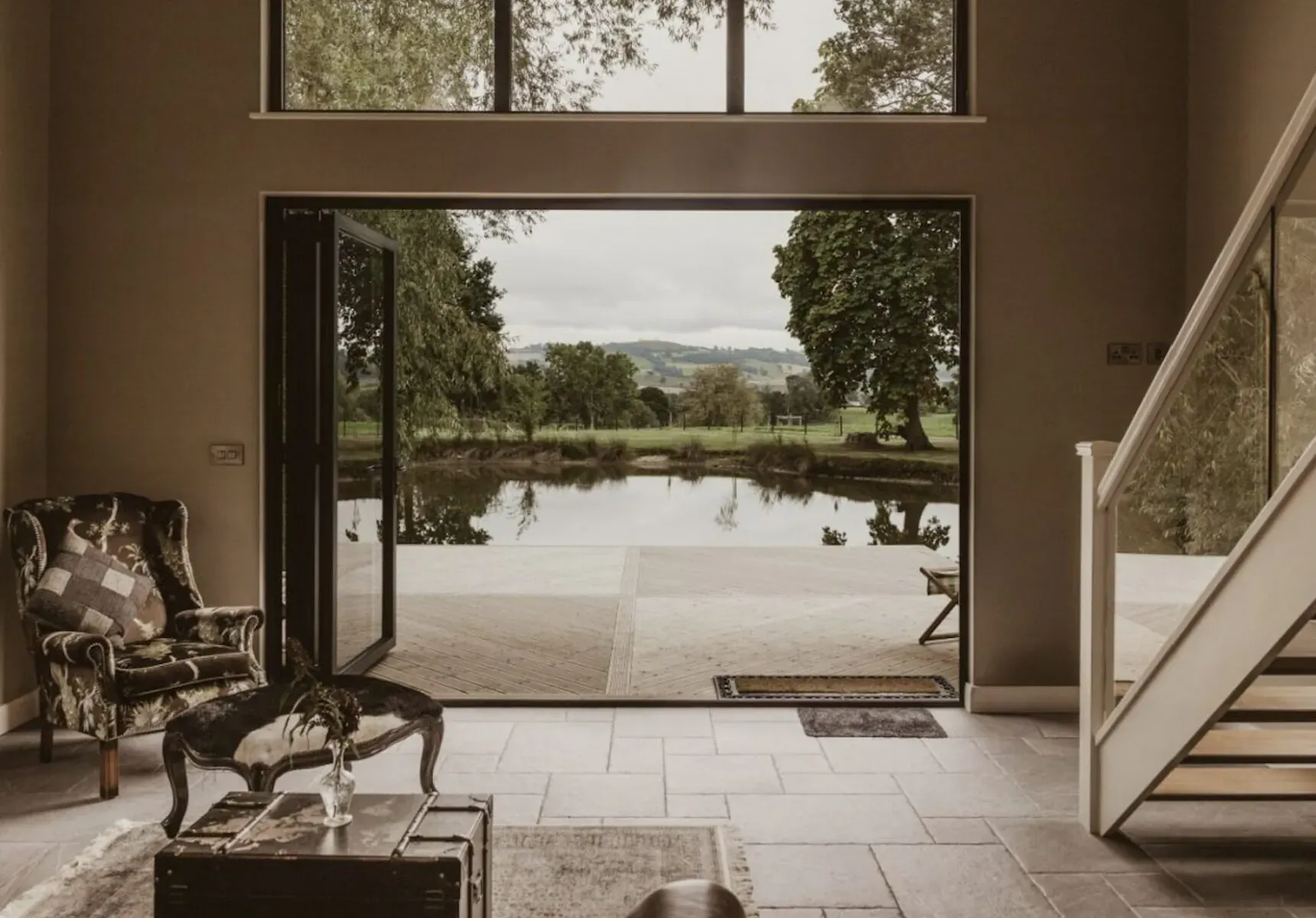 Spacious interior of The Boathouse in Wales with patterned armchair, open folding doors to private lake and hills.