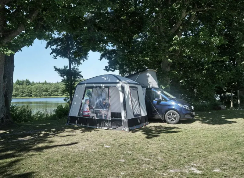 Gray luxury awning tent attached to blue campervan parked on grassy lakeside with trees, people inside.