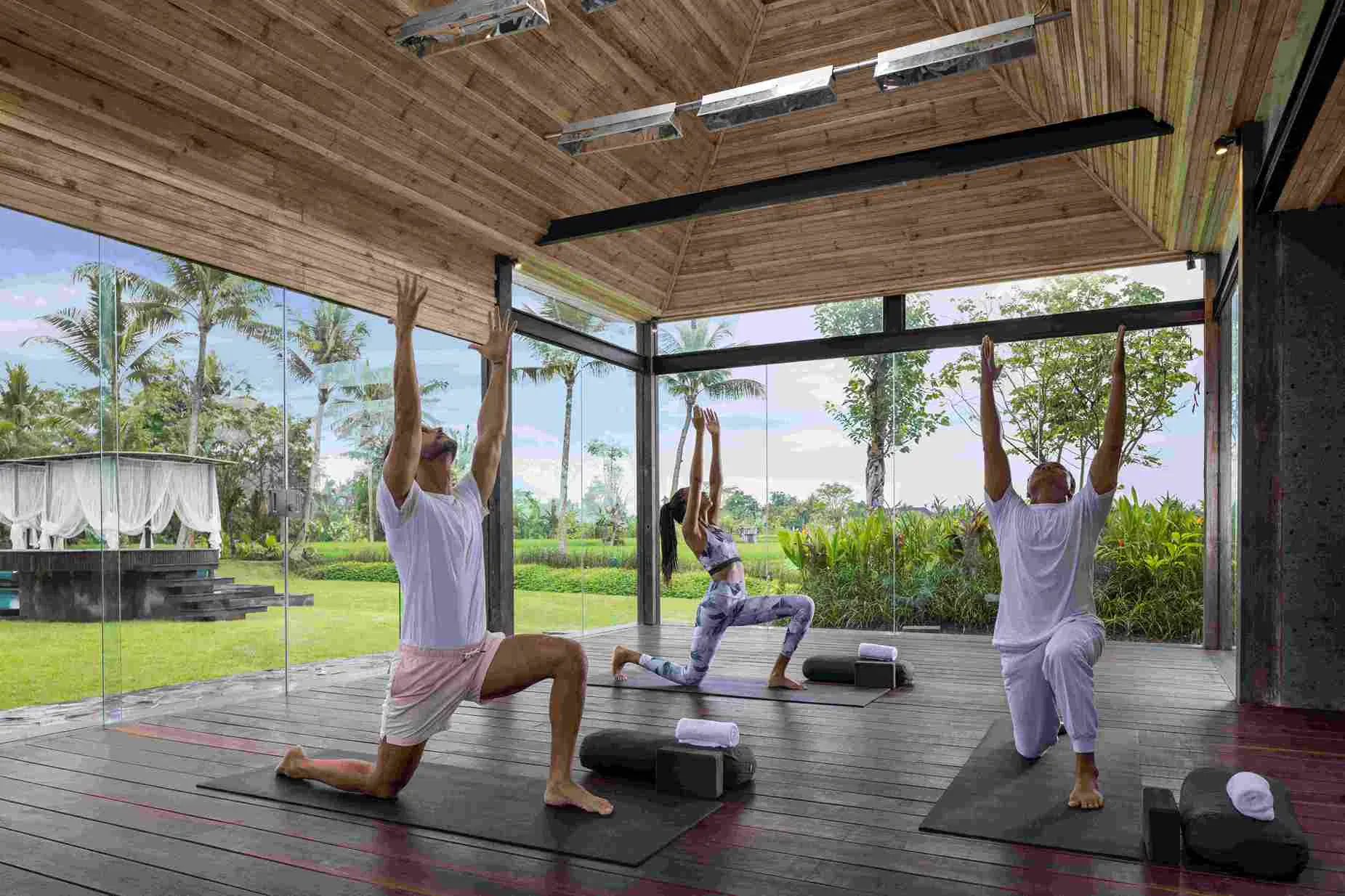 Three people practicing yoga poses with blocks on mats in open pavilion overlooking Bali garden and palms