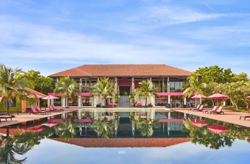 Red-roofed tropical resort building reflected in infinity pool, surrounded by palm trees, pink umbrellas, and loungers under blue sky.