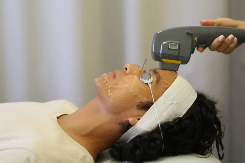 Woman lying on treatment bed with white headband and facial mask, receiving laser therapy on face from clinician in clinic.