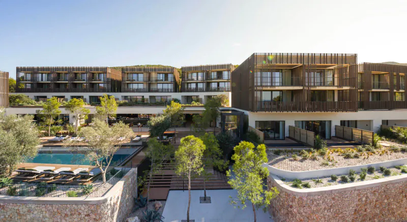 Aerial view of Maslina Resort's modern wooden buildings, lush gardens, and turquoise pool overlooking the sea.