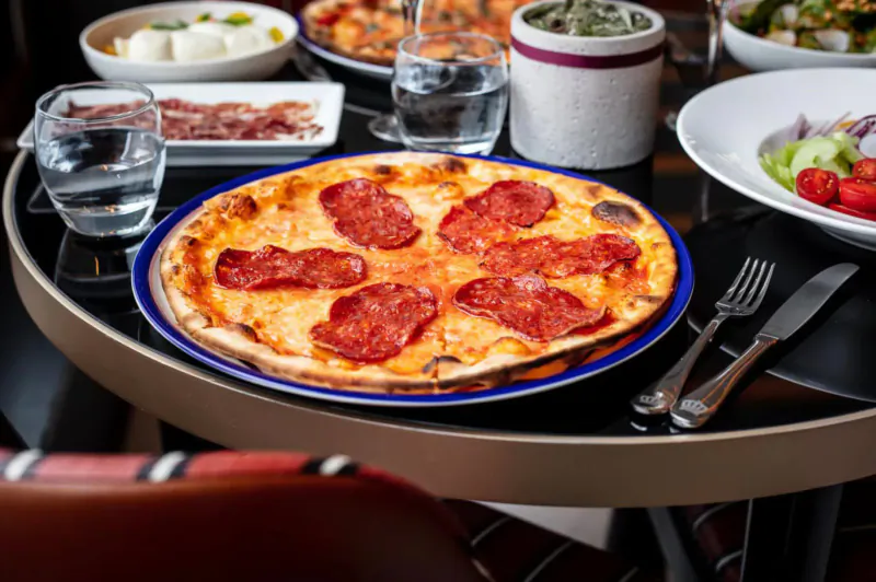 Pizza with chorizo slices on blue plate at restaurant table with salads, water glasses, and cutlery