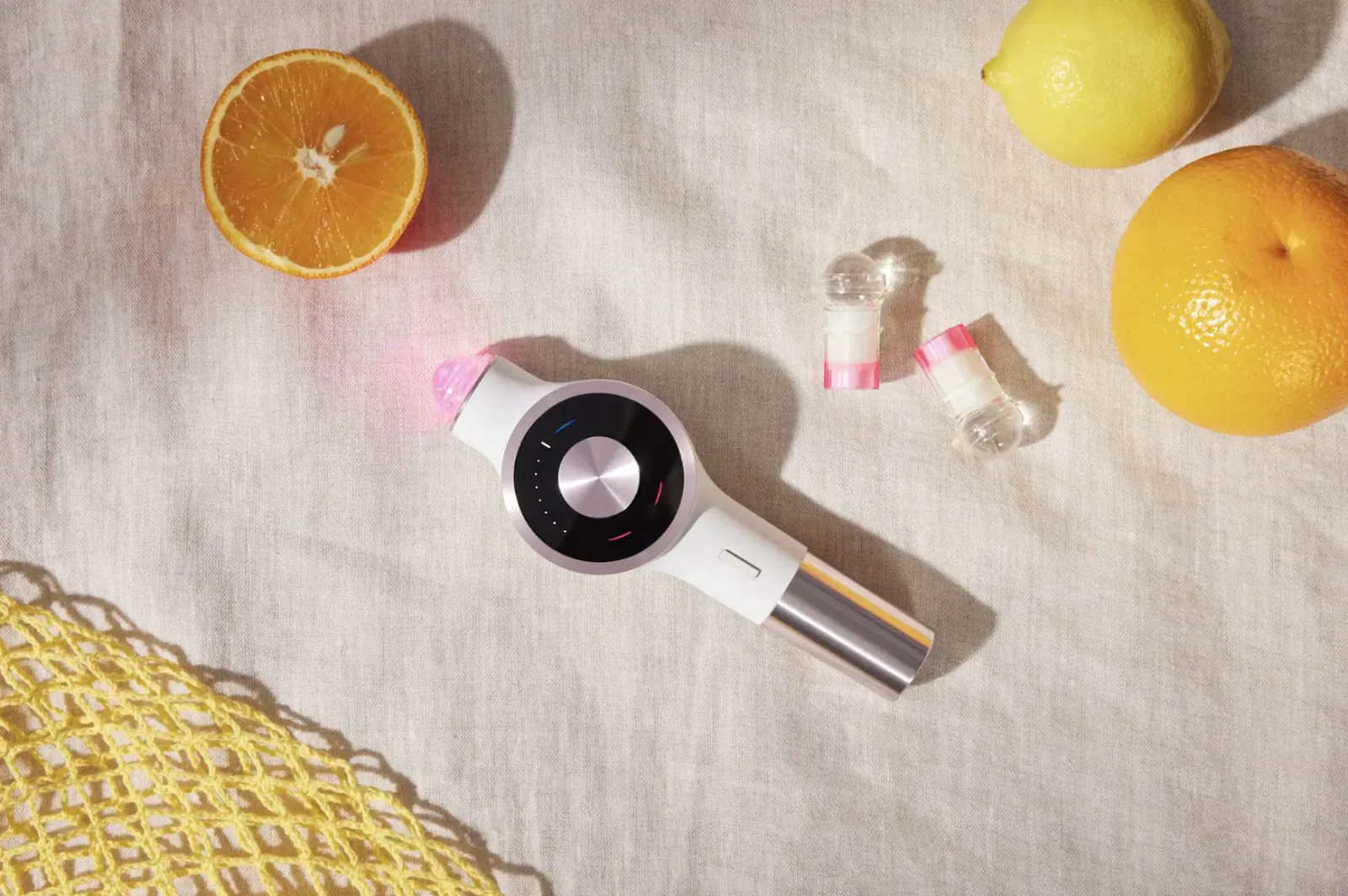 Lightinderm LED + Serum device with red light on white surface, surrounded by orange slices, lemon, earplugs, and yellow netting.