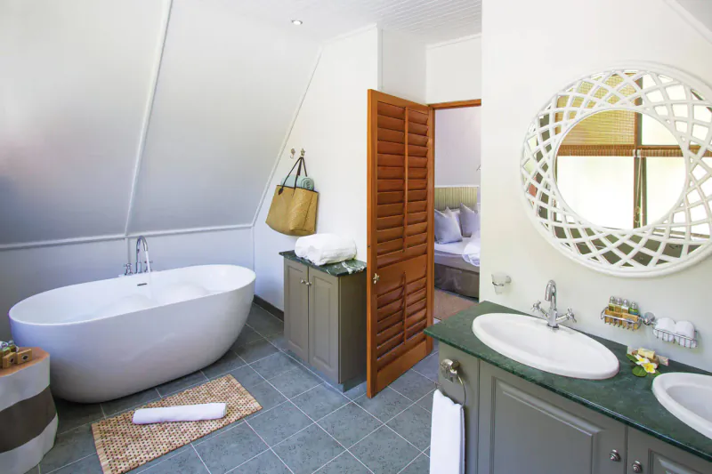 Luxurious Seychelles bathroom with freestanding tub, double sinks, teak shutters to bedroom, and woven mirror.