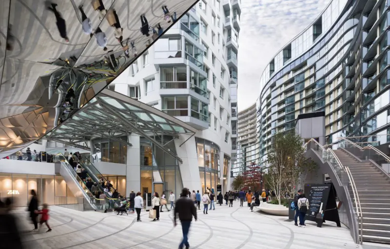 Evening view of Battersea Power Station: curved modern buildings, escalators, stairs, and people walking in riverside piazza.
