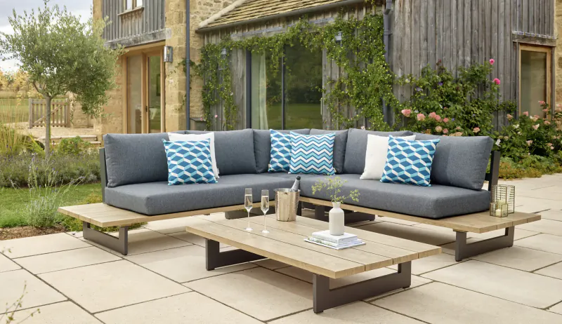 Grey L-shaped outdoor sofa with blue cushions and wooden coffee table on patio, ivy-covered stone barn backdrop.