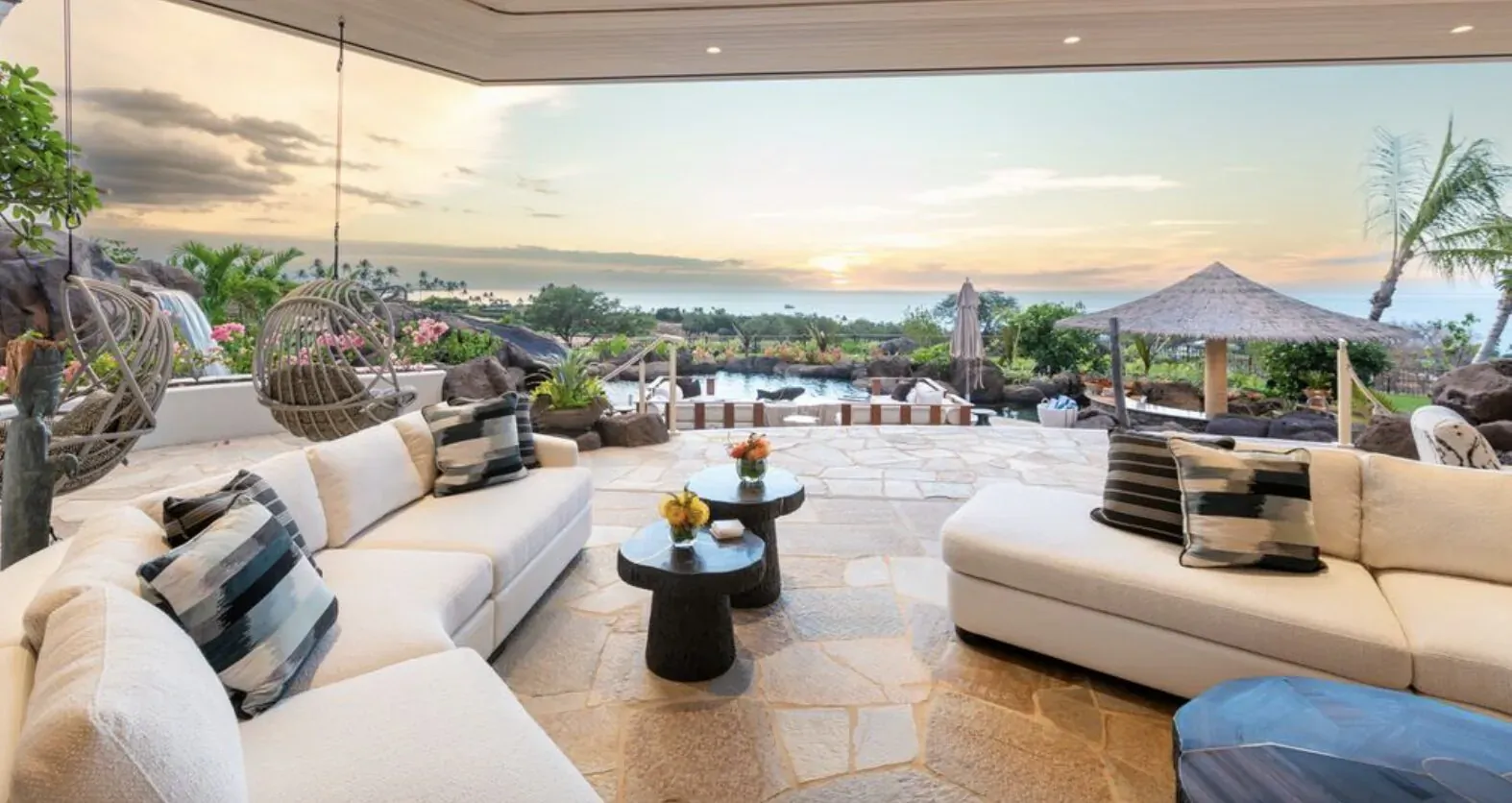 Luxurious outdoor villa living area with white sofas, coffee tables, pool, and sunset ocean view.