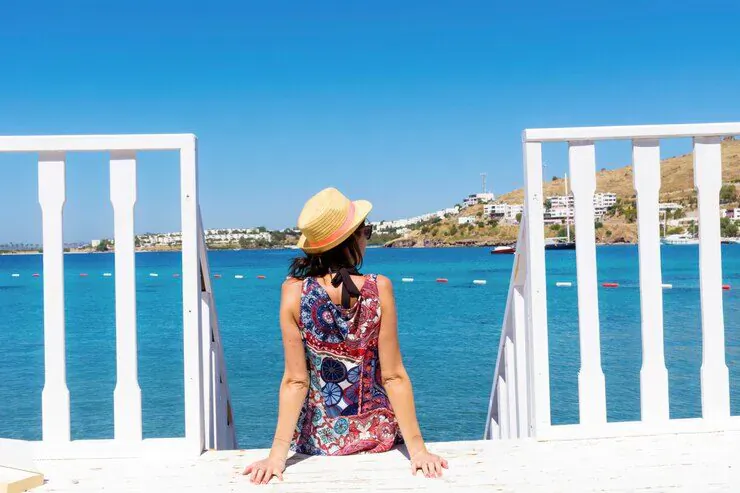 Woman in yellow hat and floral dress sits on white steps overlooking blue sea and hillside village