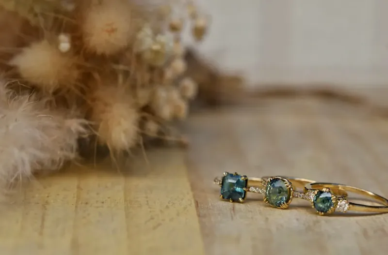 Three gold rings with green gemstones on wooden surface amid dried pampas grass.