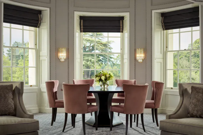 Elegant dining room at The Langley Hotel with pink upholstered chairs around black table, yellow flowers, large sash windows with blinds overlooking trees.