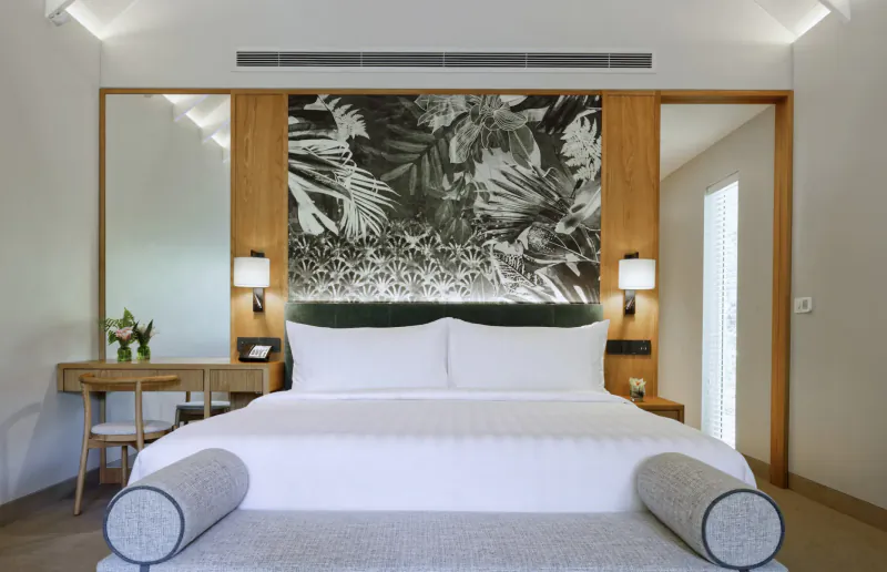 Luxurious hotel bedroom with white bed, tropical leaf wall art headboard, bedside lamps, desk, and mirror in Amari Raaya Maldives