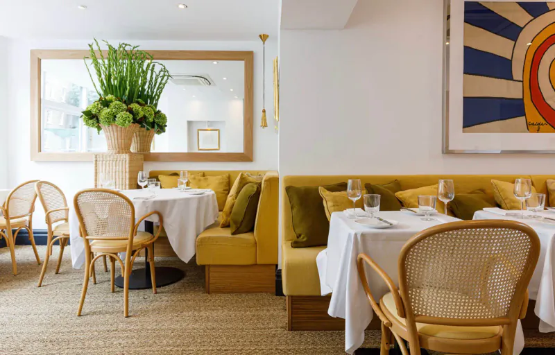 1771 Restaurant interior: rattan chairs at white tables, yellow velvet banquettes, large potted plant, abstract wall art.
