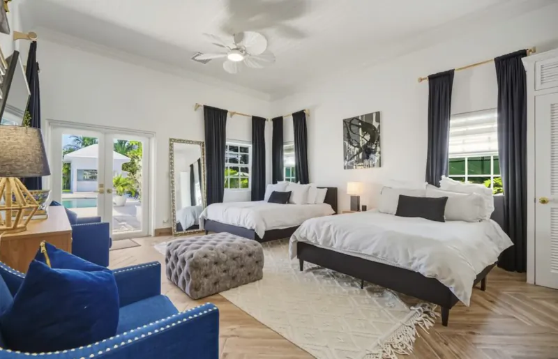 Spacious Lyford Cay bedroom with two queen beds, blue armchair, ottoman, desk lamp, pool view via French doors.