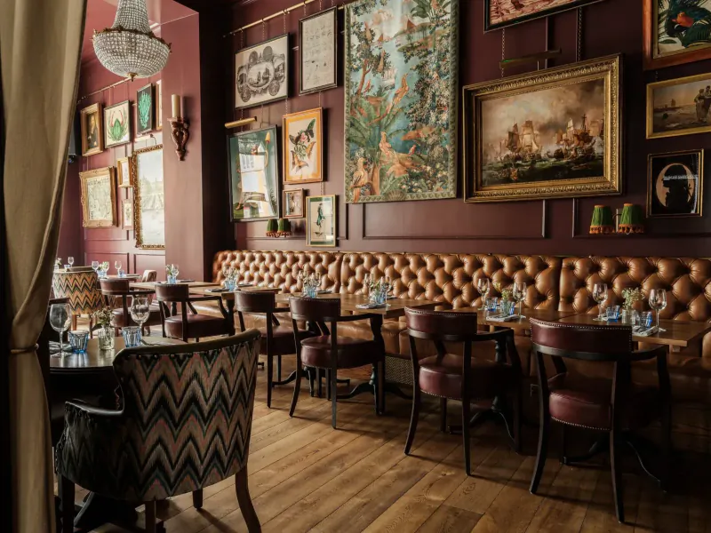 Elegant interior of The Trafalgar Public House with tufted maroon booths, wooden tables set for dining, ornate framed art on walls, and chandelier.