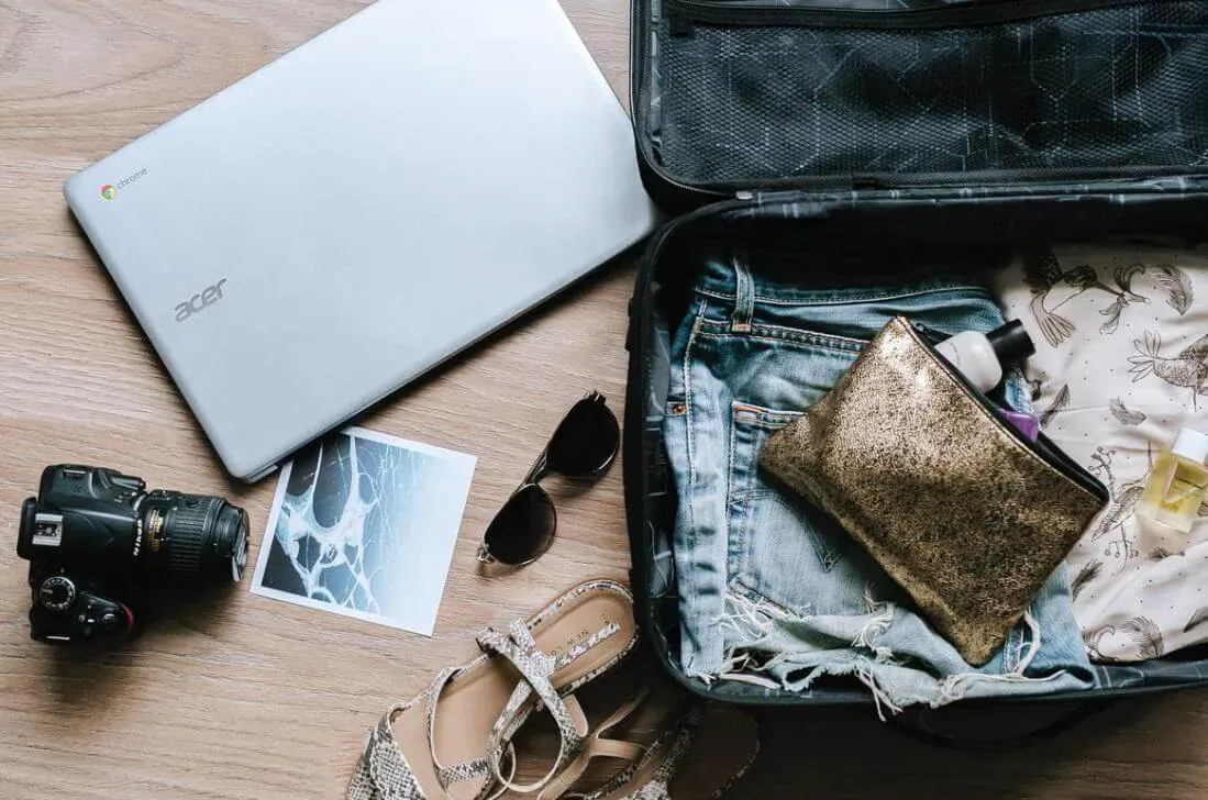 Open black suitcase with packed jeans, gold clutch, perfume, white Acer Chromebook, camera, sunglasses, sandals, and photo on wood surface