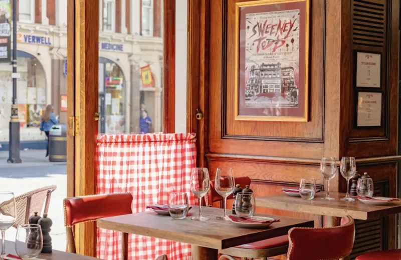Cozy Italian steakhouse interior with red checkered curtains, wooden tables set with wine glasses, and 'MACELLAIO TOPS' poster, street view outside.