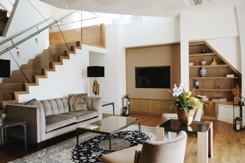 Modern luxury living room with gray tufted sofa, glass staircase, TV, orchids, and wooden decor