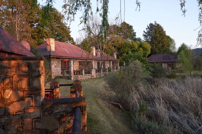 Stone cottages with red roofs at golden hour sunset, surrounded by trees, grass, and a stream.