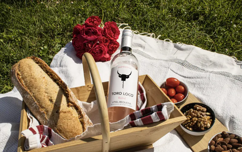 Picnic basket on grass with Nordo Loco rosé bottle, baguette, red tomatoes, nuts, red roses bouquet