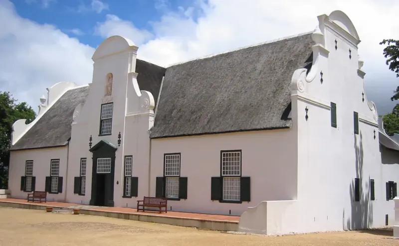 White Cape Dutch manor house with thatched roof, green shutters, benches, at Groot Constantia under blue sky.