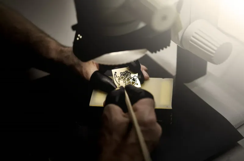 Close-up of hands using microscope and brush to detail gold BOVET Récital 28 watch dial on black workbench.