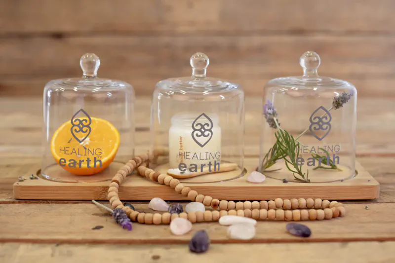 Three glass cloches on wooden tray: 'Healing Earth' logo with orange slice, candle, lavender, herbs, beads, crystals.