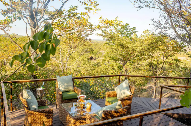 Outdoor deck at Old Drift Lodge with wicker chairs, green cushions, drinks on glass table, overlooking lush savanna landscape.