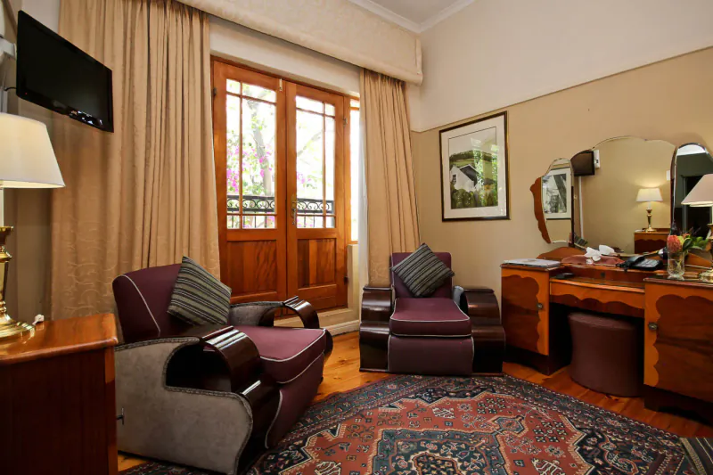 Elegant hotel room with plush armchairs, vanity table, wooden doors to balcony, and Persian rug at Montagu Country Hotel
