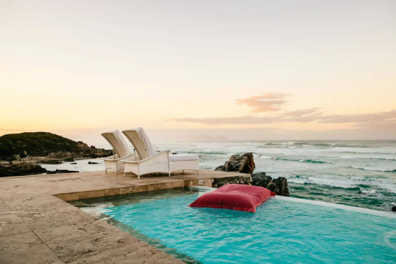 Infinity pool with red pillow and white lounge chairs on terrace overlooking ocean at sunset in Hermanus