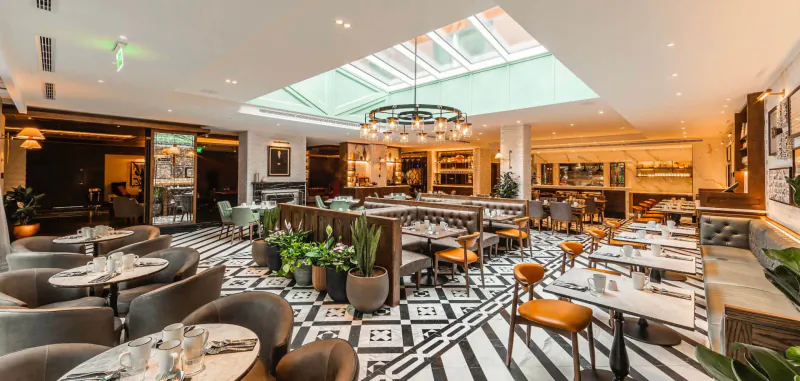 Elegant apartment-style dining room at The Fellows House Cambridge with black-and-white checkered floors, green plants, round tables, orange chairs, and skylight.