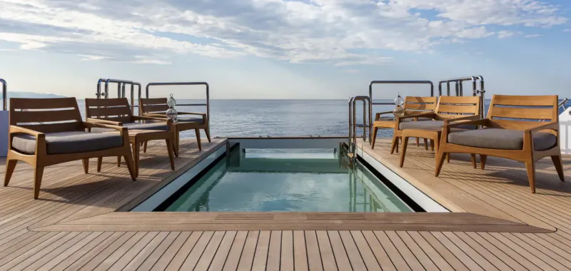 Infinity glass pool on luxury yacht deck with wooden lounge chairs, ocean horizon under partly cloudy sky